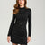 A woman wearing a fitted black long-sleeve mock-neck dress with a matching waist belt and small gold buckle, photographed against a plain light gray studio background.