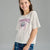 A young girl smiling while wearing a white short-sleeve graphic t-shirt with a purple and blue Tennessee Volunteers logo, paired with blue patterned shorts, against a solid gray studio background.