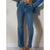 A person wearing medium-wash blue denim jeans with a straight-to-slightly-flared leg, paired with a matching blue denim shirt draped off the shoulders, standing against a plain off-white wall.