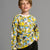 A young boy with light brown hair smiling, wearing a long-sleeve crewneck sweatshirt covered in a yellow and blue cartoon minion pattern, paired with dark jeans, against a solid gray studio background.