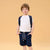 A young boy stands against a solid beige studio background wearing a white raglan t-shirt, navy blue shorts with a small white pattern, a matching navy backpack, and navy sneakers.