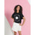 A young girl with curly dark hair stands against a pink and white geometric background, wearing a black graphic t-shirt with a white sun design and text, paired with white wrap-style shorts.