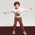 A young boy modeling a light gray sweatshirt with a brown graphic print and brown pants, posing with outstretched arms against a split white and dark brown studio background.
