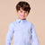 A young boy with brown hair wearing a light blue long-sleeve button-down shirt, standing against a solid beige background.