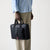 A person wearing a light blue shirt, beige trousers, and dark brown leather oxford shoes holds a black briefcase-style bag with top handles and a small red, white, and blue striped accent on the front, photographed against a clean light gray studio background.