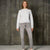 A woman stands against a textured concrete wall wearing a light gray speckled knit sweater, gray jeans, and white sneakers.