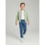 A young boy walking forward in a studio setting, wearing a light sage green zip-up bomber jacket over a white t-shirt, blue jogger pants, and white sneakers.