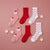 Four Christmas-themed socks arranged on a dusty pink background with small cream star decorations. The set includes two light pink socks with Santa Claus faces and two red socks, plus two white socks with red polka dots.