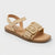 A single toddler girl's flat sandal in beige, featuring a woven jute-style double-buckle strap across the footbed and a matching ankle strap, shown on a light gray background.