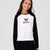 A young girl smiling and wearing a black and white long-sleeve raglan shirt with a cartoon character graphic and the word 'Anomi' on the chest, paired with a black pleated skirt, against a light gray studio background.