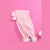 A light pink knit accessory, likely a hat or scarf, laid flat on a solid pink background with two white pom-poms and fluffy pastel trim at the ends.