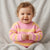 A smiling baby wearing a pink and yellow striped knit sweater, sitting on a soft gray blanket.