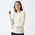 A woman modeling a cream-colored, long-sleeve crewneck knit sweater against a plain white studio background.