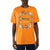 A man wearing a bright orange short-sleeve t-shirt with a circular floral wreath graphic on the chest featuring the words 'Agath Tennis'.