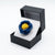 A blue and yellow fabric flower brooch displayed inside an open black presentation box with a white insert, set against a plain white background.