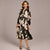 A woman modeling a long-sleeve midi dress with a black and beige abstract floral print, featuring a tie-neck detail and a self-tie belt, set against a solid gray studio background.