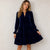 A woman wearing a navy blue velvet tiered dress with long sleeves and a split neckline, standing against a textured off-white wall.