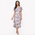 A woman modeling a short-sleeved, tiered midi dress with a watercolor floral print in purple, pink, and green on a white base, set against a plain white background.