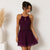 A woman modeling a sleeveless halter mini dress in deep burgundy purple. The dress features a lace-embroidered bodice with a keyhole cutout and a flowing chiffon skirt, set against a neutral white wall with a decorative vase of pampas grass on the left.