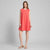A woman modeling a sleeveless coral pink shift dress with a round neckline and a flared silhouette, paired with tan heeled sandals, against a light gray studio background.