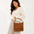 A woman wearing a cream knit sweater and light beige pants holds a brown shoulder bag with a top handle and a decorative stitched band near the bottom. The brand name 'SOL'S' is visible in gold lettering on the front of the bag.