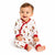 A smiling baby sitting on a white studio background wearing a white one-piece pajama with red trim and a festive holiday print.