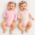 Two smiling babies lying side by side on a white textured blanket. The baby on the left wears a solid light pink long-sleeve bodysuit and a pink bow headband.