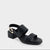 A single black leather sandal with a low block heel, double front straps, and a gold-tone chain detail on the ankle strap, shown against a light gray background.
