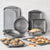 A 15-piece non-stick baking pan set in dark gray, displayed on a white countertop against a white subway tile backsplash. The set includes various rectangular, round, and square pans, a muffin tin, and a pizza pan, with some baked goods (muffins, cookies, pizza) shown in use.