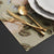 Gold-colored flatware, including a fork, knife, and spoon, arranged on a beige floral placemat next to white plates.