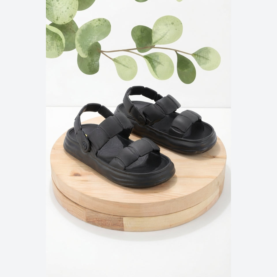 A pair of black double-strap platform sandals displayed on a round wooden riser against a white background with a decorative green leafy branch at the top.