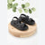A pair of black double-strap platform sandals displayed on a round wooden riser against a white background with a decorative green leafy branch at the top.