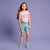 A young girl stands against a solid lavender studio background wearing a light pink sleeveless tank top with a peace sign graphic and the text 'IT'S NICE TO', light blue denim shorts, and light blue flat shoes.