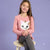 A young girl with long brown hair sits on a stool, wearing a pink long-sleeve shirt with a sequined cat face graphic and dark gray leggings against a solid lavender background.