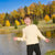 A young boy smiling and posing outdoors by a lake, wearing a cream-colored cable-knit sweater and dark pants, with a background of autumn trees and blue sky.