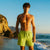 A shirtless man standing on a beach wearing bright lime green swim trunks, with ocean waves, rocks, and a clear blue sky in the background.