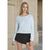 A woman modeling a light blue long-sleeve knit top with a wide scoop neckline, paired with a black mini skirt, set against a beige stone building background.