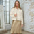 A woman models a cream-colored cropped jacket with flap pockets over a white top and a beige satin midi skirt, standing in front of a white brick wall.