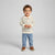 A toddler standing against a light gray studio background, wearing a cream and light gray striped long-sleeve polo shirt, light blue jeans, and brown shoes.