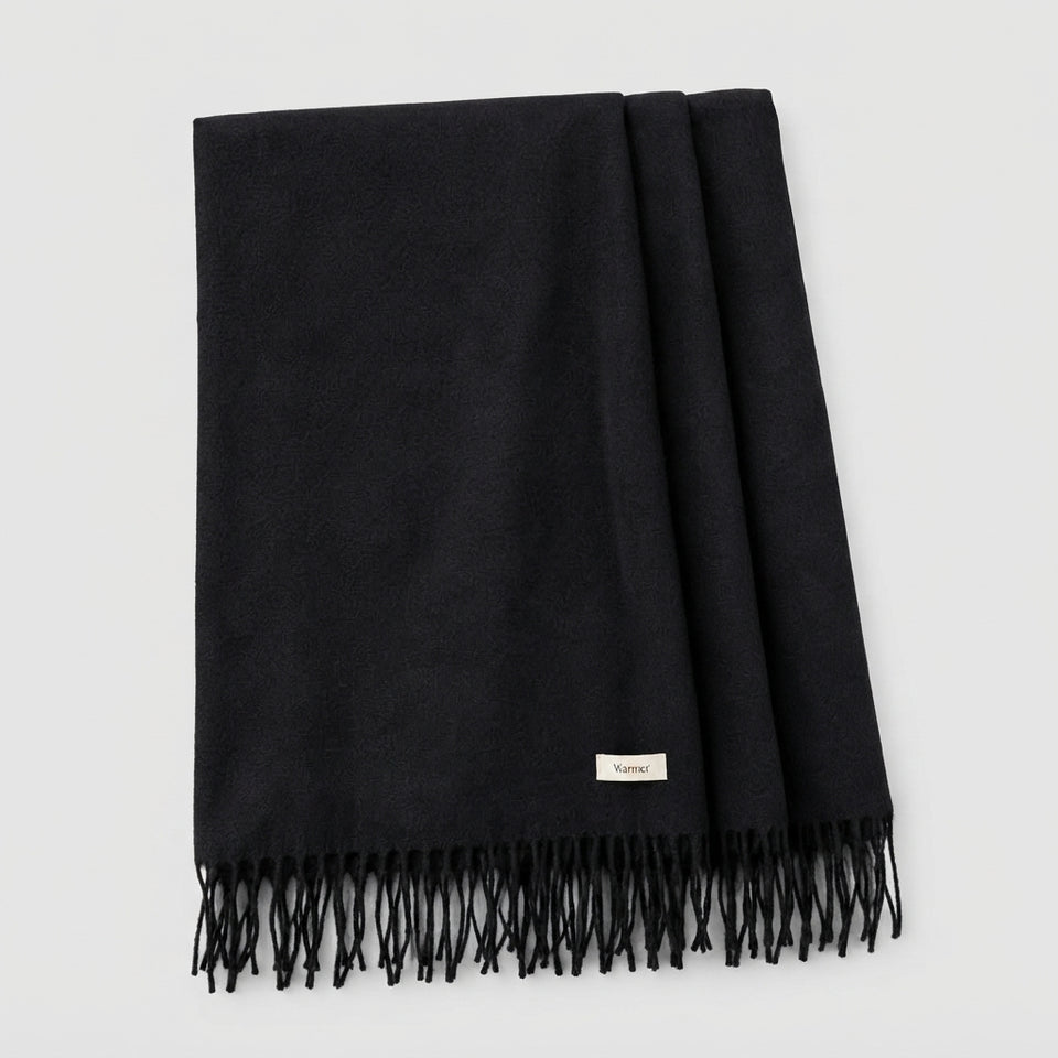A folded black woven scarf with fringe detailing and a small beige brand label, displayed on a light gray background.