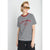 A woman modeling a black and white horizontally striped t-shirt with red trim at the collar and red embroidered text reading 'V.GOL VERNON 1963' on the front, paired with black pants and orange-tinted sunglasses against a light gray background.
