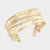 A gold wrap cuff bracelet with multiple textured bands and rows of clear rhinestones, shown on a light gray background. The brand name 'SARABACO' is engraved on the small hanging tag.