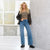 A blonde woman stands in a bright studio wearing a black leather jacket, a leopard-print strapless crop top, and high-waisted blue flare jeans with black platform boots.