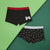 Two pairs of men's boxer briefs laid flat on a split green background. The top pair is black with a red waistband and a small white video game controller graphic with a red hat on the right side.