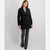 A woman modeling a black double-breasted coat with notched lapels, paired with dark gray trousers and black shoes, against a light gray studio background.