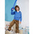 A young boy wearing a blue zip-up hoodie and brown pants stands on a beige chair in a bedroom setting, holding a blue balloon sword.