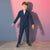A young boy modeling a navy blue two-piece suit with a white t-shirt and navy sneakers, standing against a pink and blue gradient studio backdrop.