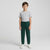 A young boy stands against a light gray studio background wearing a light gray crewneck t-shirt, dark green jogger-style pants, and white sneakers.