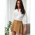 A woman wearing a white button-up shirt and a tan pleated mini skirt, standing in a bright indoor setting with a sofa and a plant visible in the background.