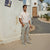 A man stands outdoors in a sunny, Mediterranean-style village setting wearing a white short-sleeve polo shirt, light gray cargo pants, and gray slip-on shoes.